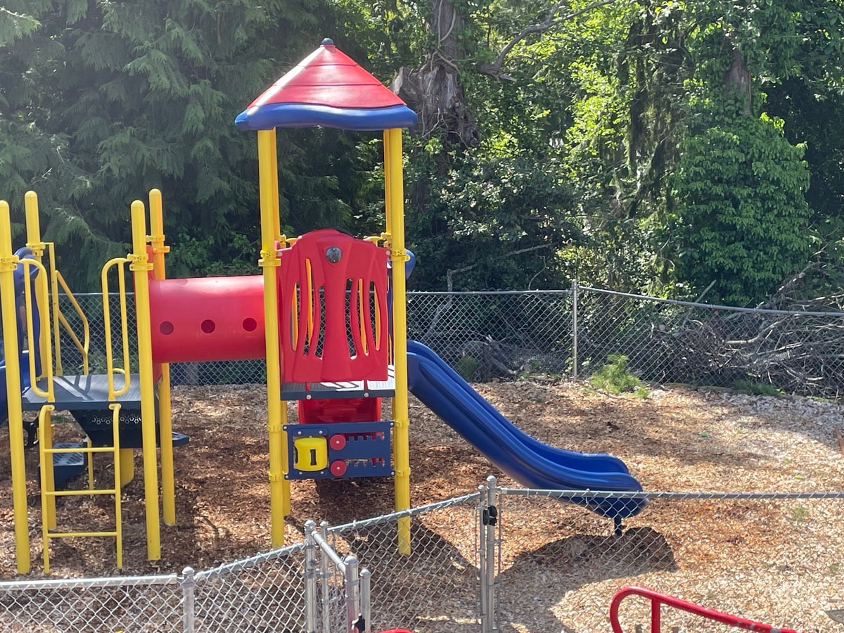 Installed red, blue and yellow playground structure on wood chips inside a chain-link fence.