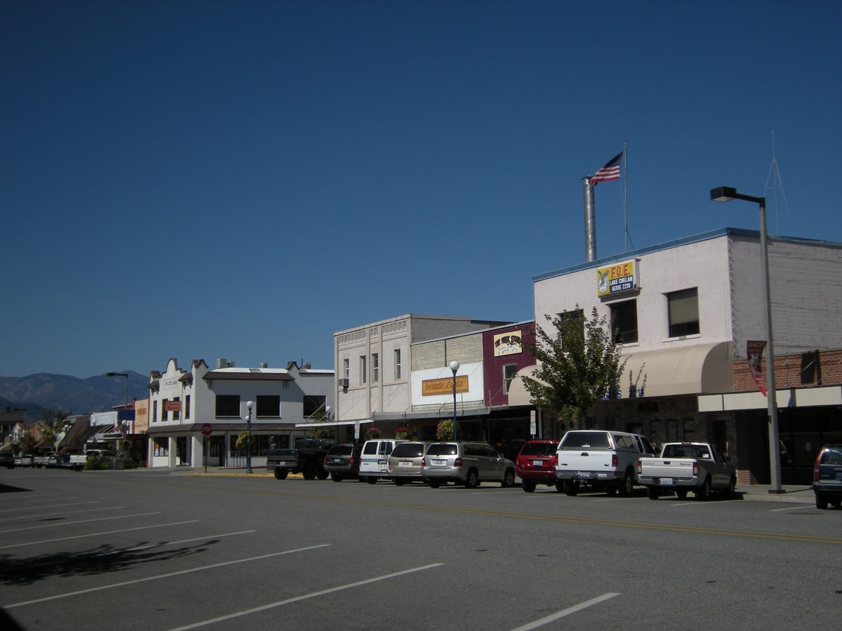 Downtown Chelan, Washington: a row of one and two storey brick and stucco storefronts along Woodin Avenue, with pickup trucks and SUVs angle parked at the kerb, an American flag on a rooftop pole, and dry brown hills rising behind the town under a clear blue sky.