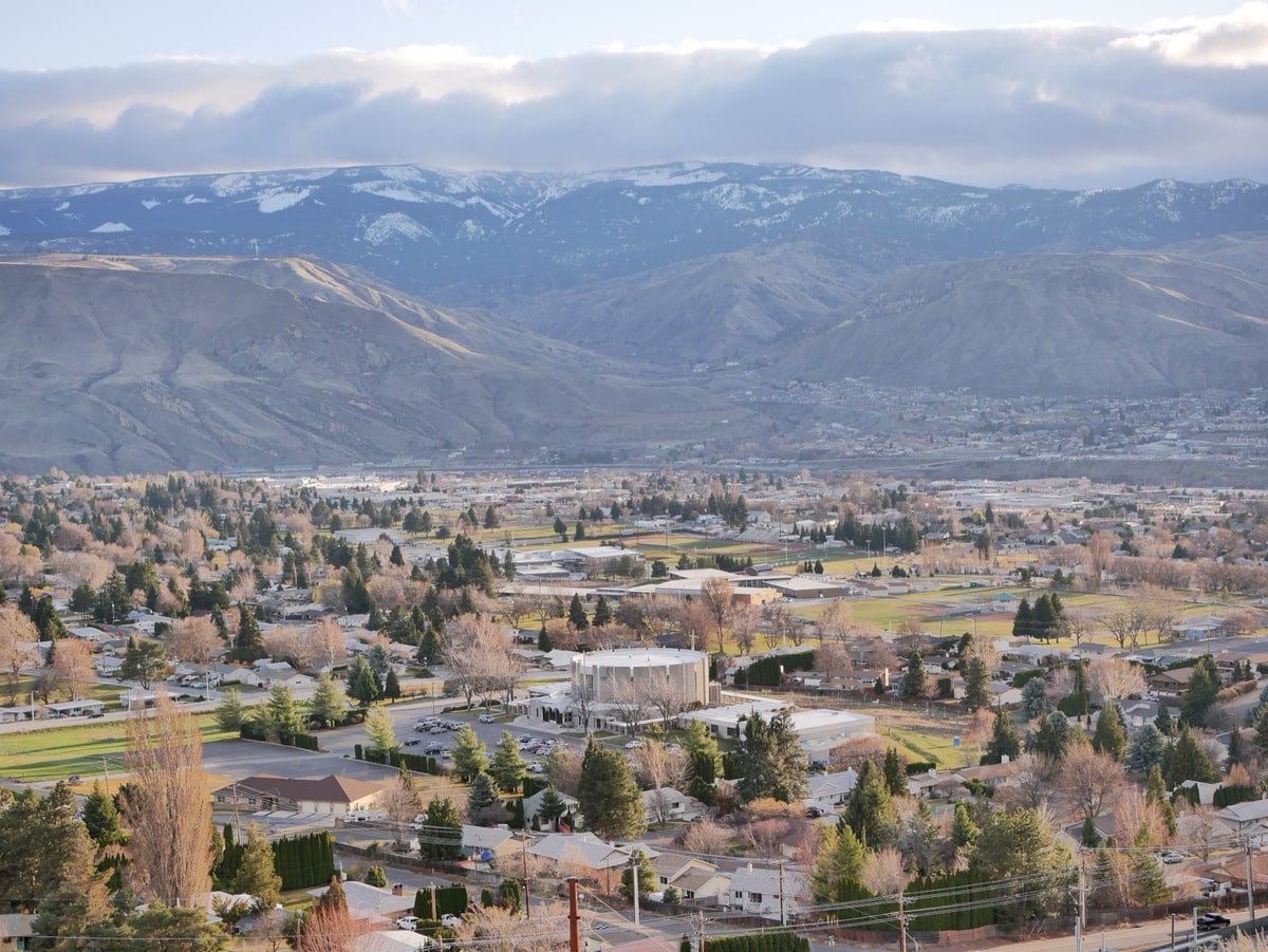 East Wenatchee, Washington seen from the hillside above the town looking south: residential streets and houses among bare early-spring trees, a school campus with running track and sports fields, a church with a rounded tower and parking lot, a commercial strip beyond, and the Columbia River valley with dry foothills and snow-dusted Cascade ridges in the distance.