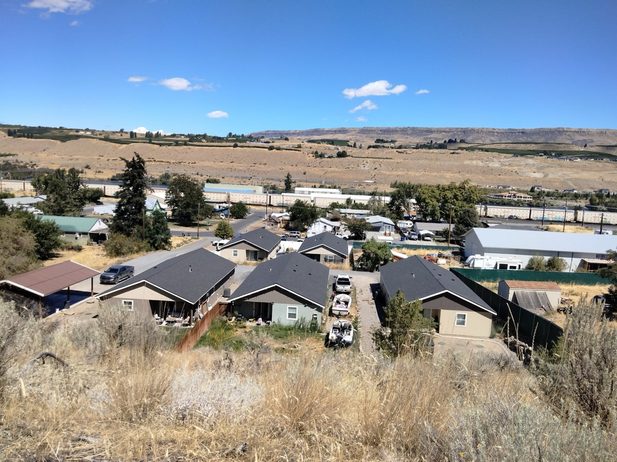 A view over Malaga, Washington from a sagebrush hillside above the community: a row of single-story houses with dark shingle roofs, fenced yards, driveways with parked pickups and boats, a white metal-sided warehouse, and a street running past.