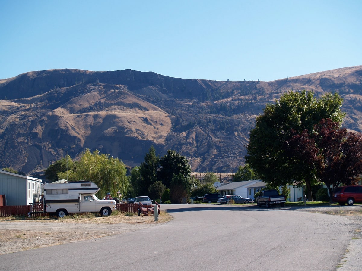 A residential street in Rock Island, Washington, lined with single storey homes, a parked pickup carrying a camper and mature shade trees, with the dry basalt bluffs above the Columbia River valley rising behind the town.