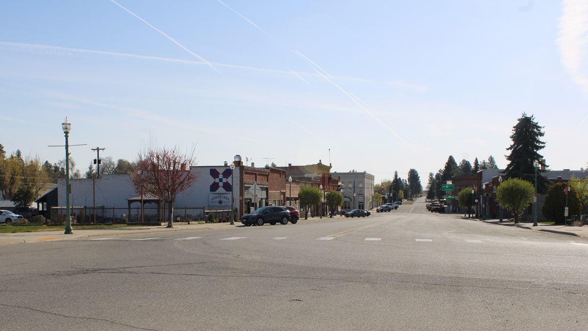 Looking down Locust Street through downtown Waterville, Washington, with early 1900s brick and painted storefronts lining both sides of the wide main street, green lamp posts, young trees and parked cars, under a clear sky streaked with contrails.